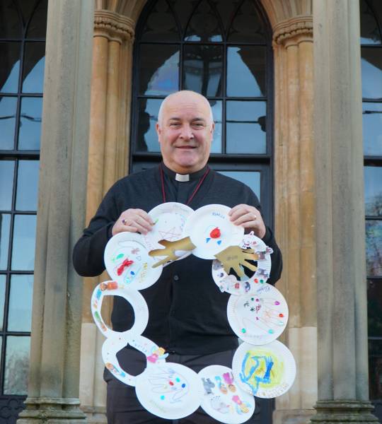 Mand standing outside holding a wreath made from card with children's drawings on it
