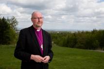 Man in black jacket and purple shirt and clerical collar standing outside with countryside behind him