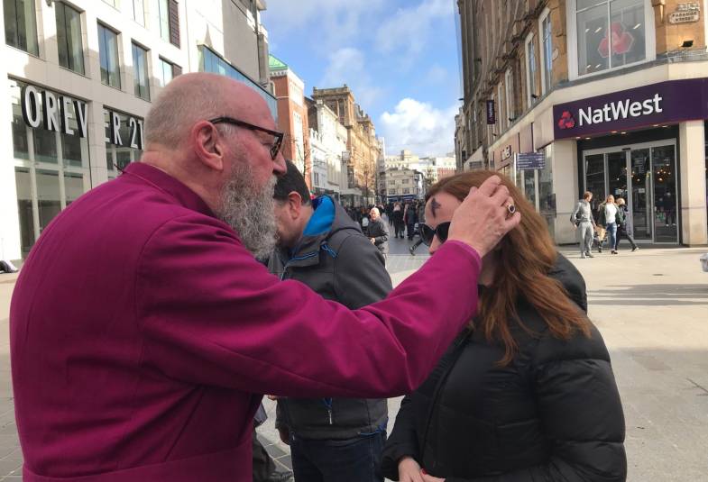 Man in purple cassock adding an ash cross to the forehead of a woman on a high street