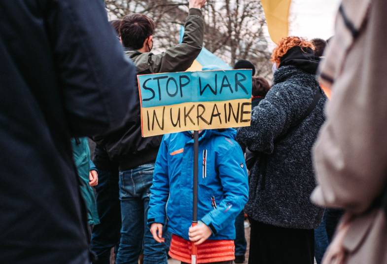 People standing outside holding flags and banners. A child's banner reads Stop War in Ukraine