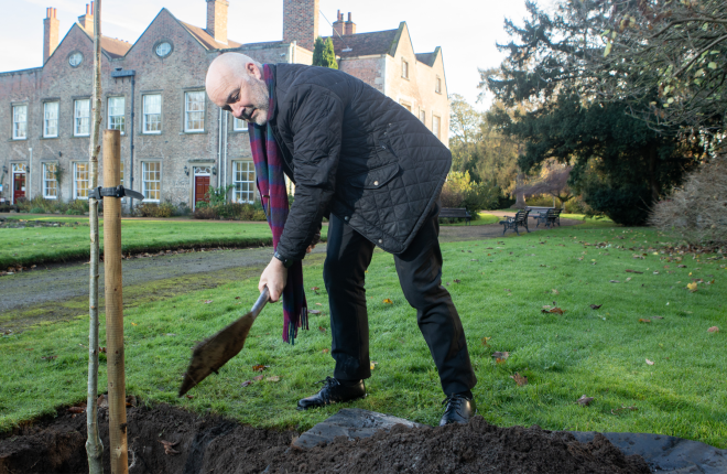 Man dressed in black using a spade to put soil around a newly planted tree 