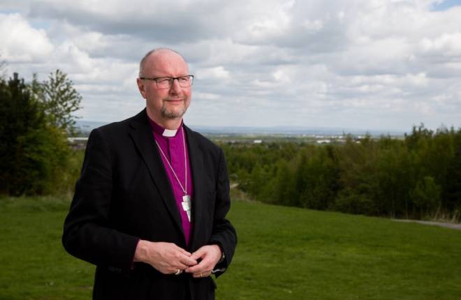 Man in black jacket and purple shirt and clerical collar standing outside with countryside behind him