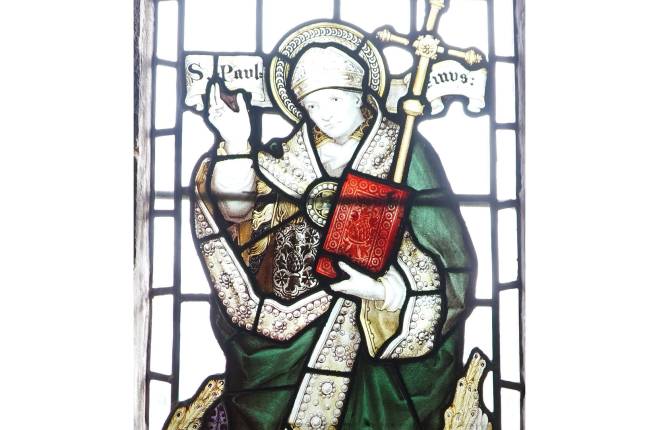 Stained glass window of St Paulinus holding bible and staff with one arm raised in blessing