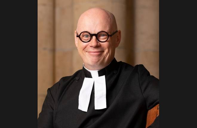 Smiling picture of Caucasian man wearing round black glasses and dressed in black cassock and dog collar